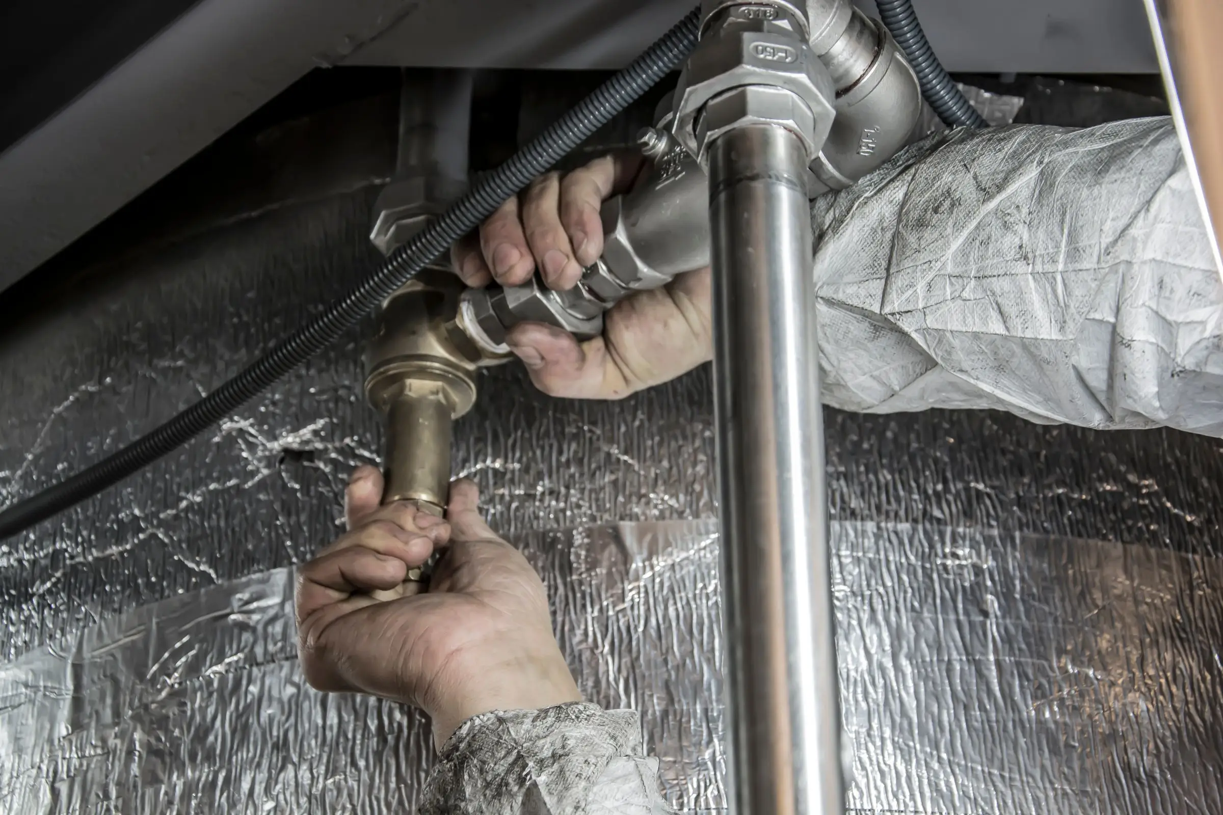 Two hands tightening a brass fitting on a pipe, foil insulation behind.