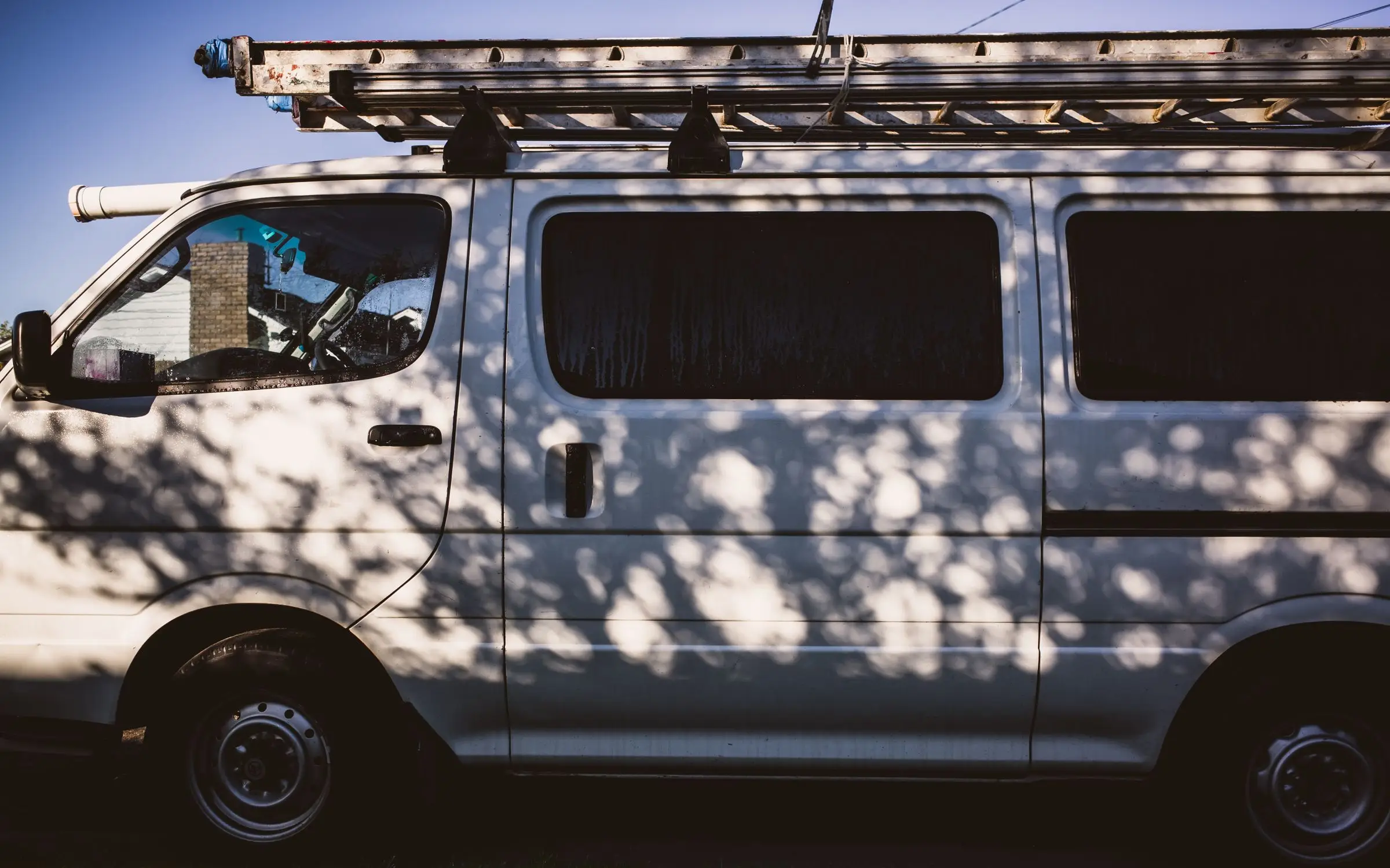A white work van with ladders on its roof rack, parked in the shade of a tree.